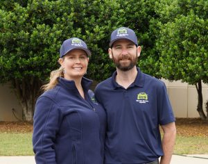 Two people in matching blue uniforms and hats stand outdoors, smiling in front of green trees.