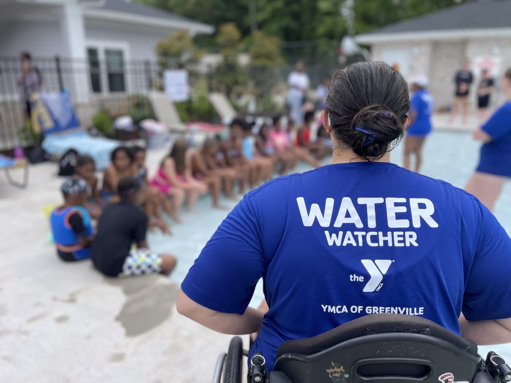 A person in a "Water Watcher" shirt oversees children sitting by a pool at a YMCA event.