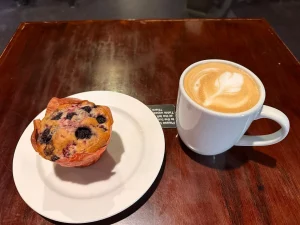 A blueberry muffin on a plate and a cup of latte with foam art on a wooden table.