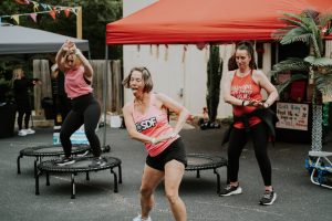 Three women in athletic wear dancing and exercising on mini trampolines outdoors under red and gray tents.
