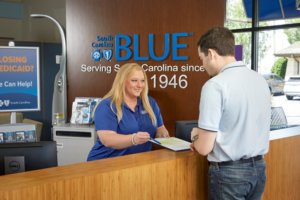A woman at a South Carolina Blue desk hands paperwork to a man standing on the customer side of the counter.