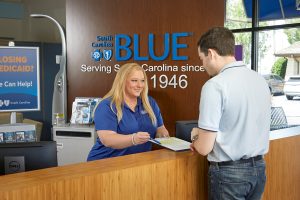 A woman at a South Carolina Blue desk hands paperwork to a man standing on the customer side of the counter.