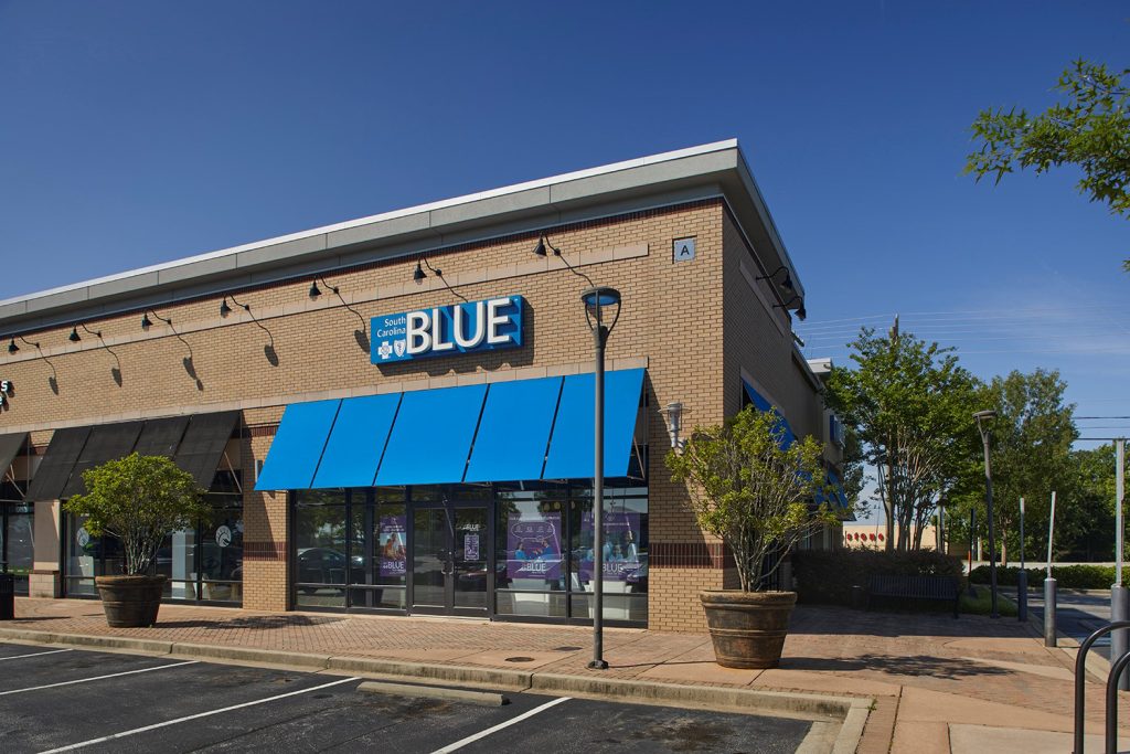 A brick storefront with blue awnings and a "BLUE" sign, next to an empty parking lot on a sunny day.
