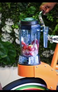 A blender with fruit sits on a bike, powered by pedaling, with greenery in the background.
