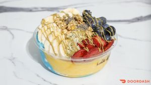 A smoothie bowl topped with banana, strawberries, blueberries, granola, seeds, and a drizzle of sauce.