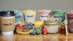 Assorted colorful smoothies, a coffee cup, yogurt jars, fresh fruit, and honey on a wooden table.