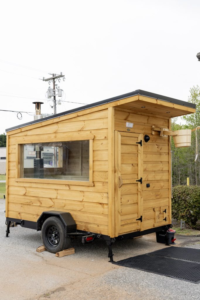 A small wooden tiny house on wheels parked outdoors with a large window and a slanted roof.