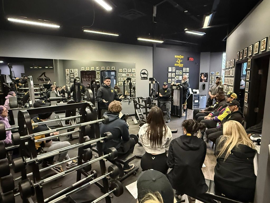 A group of people sit and listen to a speaker in a gym filled with exercise equipment and wall photos.