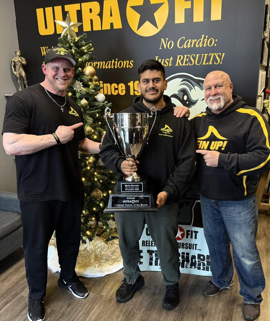 Three men smile and pose with a large trophy in front of a Christmas tree and an Ultra Fit sign.