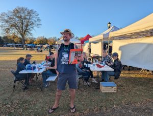 A man in casual clothes holds a clipboard, standing outdoors near tents and people sitting at tables.