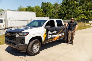 Man stands next to a Chevrolet truck with "Power Champions" branding parked on a sunny pavement.