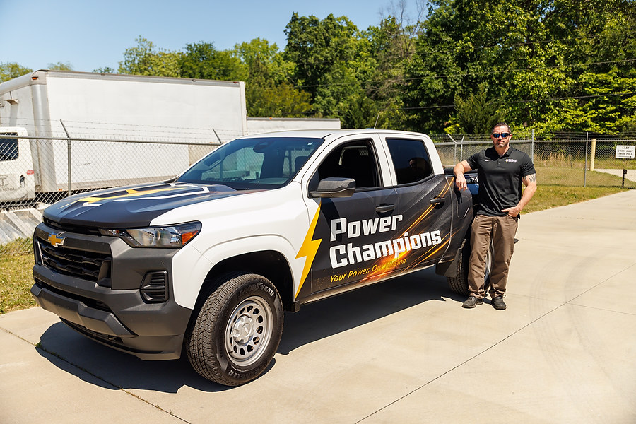 Man stands next to a Chevrolet truck with "Power Champions" branding parked on a sunny pavement.