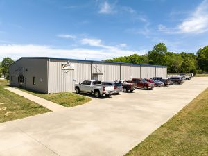 A metal industrial building with cars parked outside on a sunny day, surrounded by trees and clear skies.