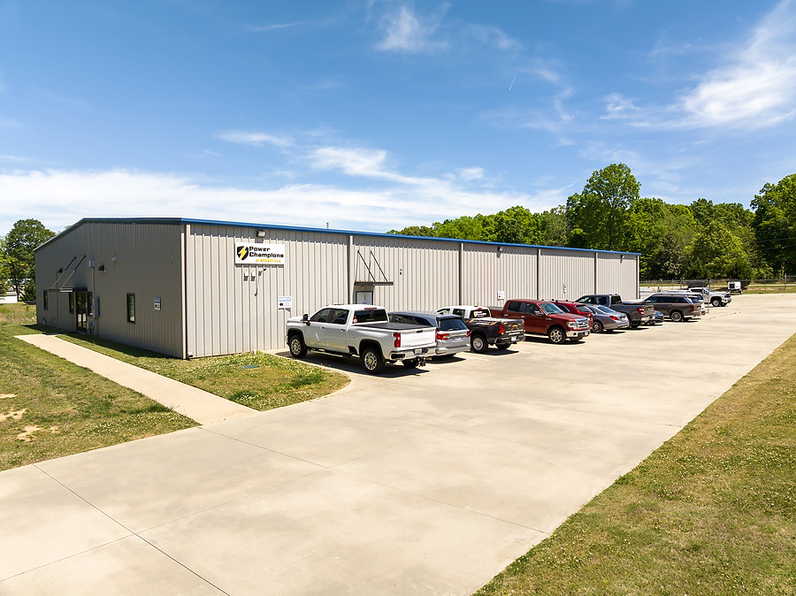 A metal industrial building with cars parked outside on a sunny day, surrounded by trees and clear skies.