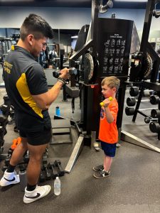 A trainer coaches a young boy lifting dumbbells at a gym, both smiling and facing each other.