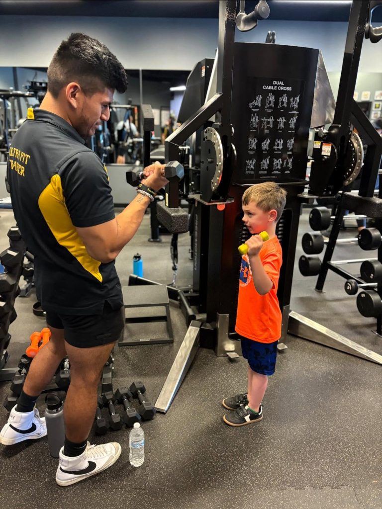A trainer coaches a young boy lifting dumbbells at a gym, both smiling and facing each other.