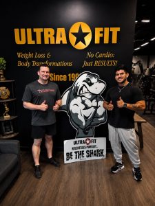 Two men posing with thumbs up in front of an "Ultra Fit" gym sign and a cartoon shark cutout.