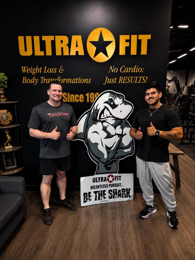 Two men posing with thumbs up in front of an "Ultra Fit" gym sign and a cartoon shark cutout.