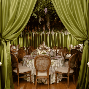 Elegant dinner tables with floral decor and wooden chairs behind green curtains and hanging greenery.