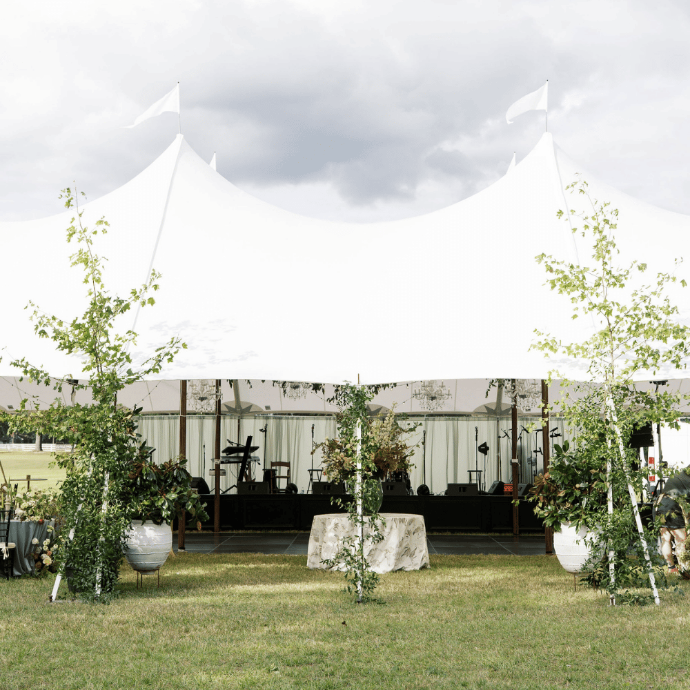 White event tent with greenery and potted plants on grass, under a cloudy sky.