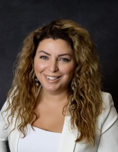 Smiling woman with long curly hair wearing a white blazer, posing in front of a dark background.