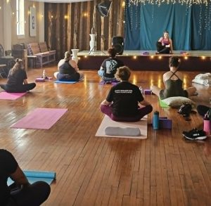 A group yoga class sits on mats in a studio with wood floors and string lights, listening to the instructor.