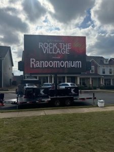 Large outdoor screen displays "ROCK THE VILLAGE presents Randomonium" in a suburban neighborhood.