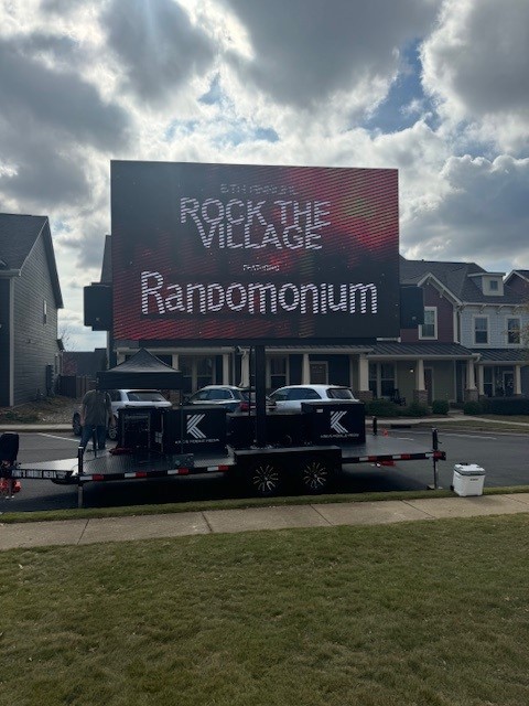 Large outdoor screen displays "ROCK THE VILLAGE presents Randomonium" in a suburban neighborhood.