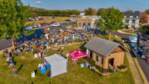 A large crowd gathers on a lawn for an outdoor event near small buildings and tents on a sunny day.