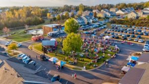 A large crowd gathers at a neighborhood park event surrounded by houses, trees, tents, and parked cars.