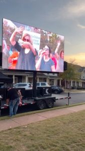 A man stands by a trailer as a large outdoor screen shows people smiling and waving at an event.