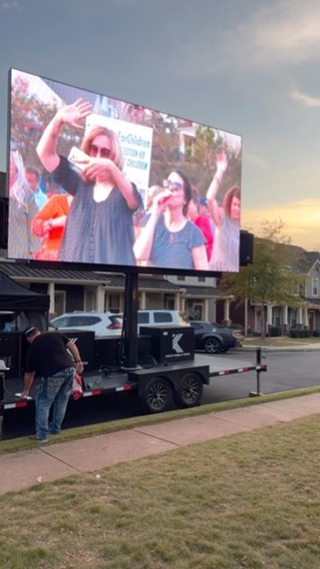 A man stands by a trailer as a large outdoor screen shows people smiling and waving at an event.