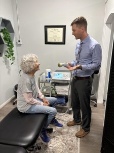 A doctor speaks to an older woman seated in a clinic, holding a medical device in his hand.