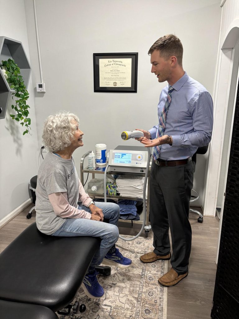 A doctor speaks to an older woman seated in a clinic, holding a medical device in his hand.