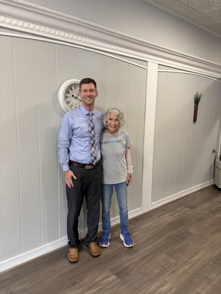 A man in business attire and an older woman in a t-shirt and jeans stand smiling in an office-like space.