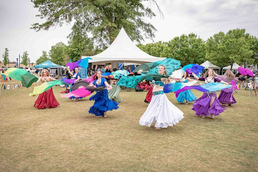A group of women in colorful skirts perform a dance with scarves outdoors at a festival.