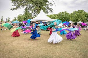 A group of women in colorful skirts perform a dance with scarves outdoors at a festival.