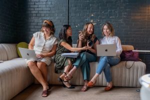 Four women sit on a couch with laptops, smiling and looking at a camera one of them is holding, as they collaborate on creative content for social media.