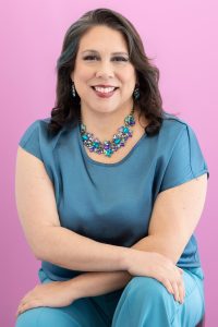 Smiling woman in blue outfit and colorful necklace sitting in front of a pink background.