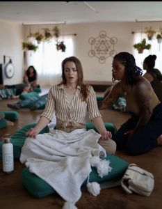 A woman meditates on a cushion with eyes closed as another woman gently guides her in a calm room.