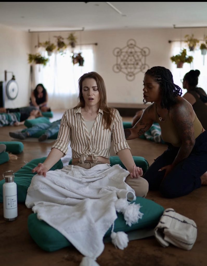 A woman meditates on a cushion with eyes closed as another woman gently guides her in a calm room.