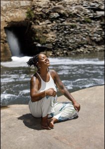 A woman sits on a rock by a river, smiling with eyes closed, enjoying the sun and nature.