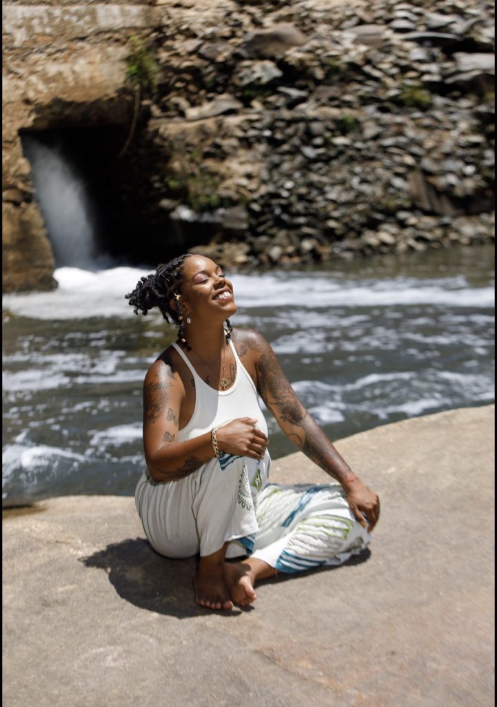 A woman sits on a rock by a river, smiling with eyes closed, enjoying the sun and nature.