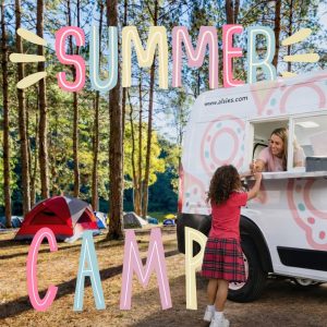A girl gets ice cream from a van at a forest campsite; text reads "SUMMER CAMP.