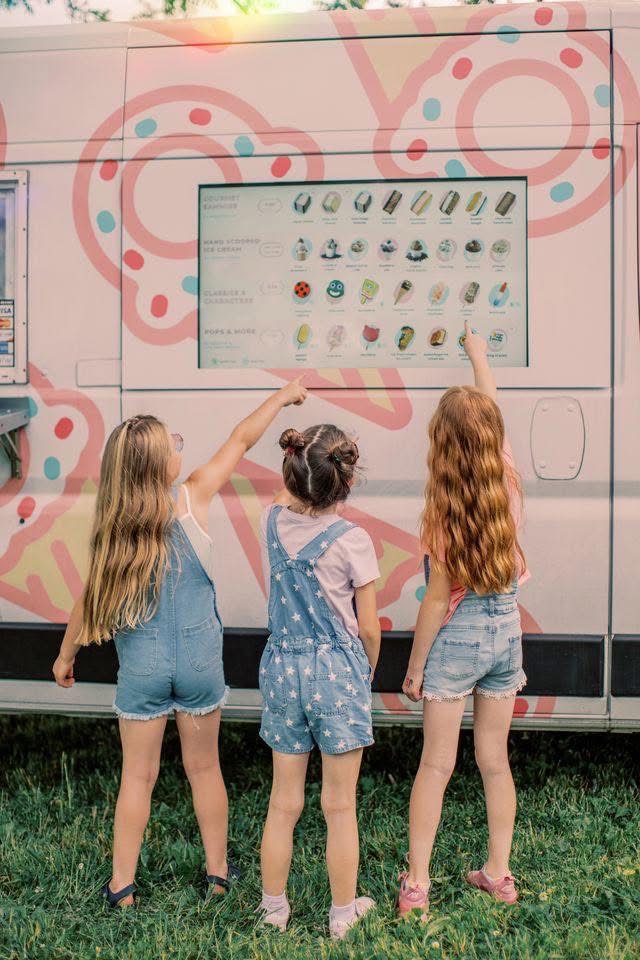 Three young girls point at the menu on the side of an ice cream truck parked on grass.