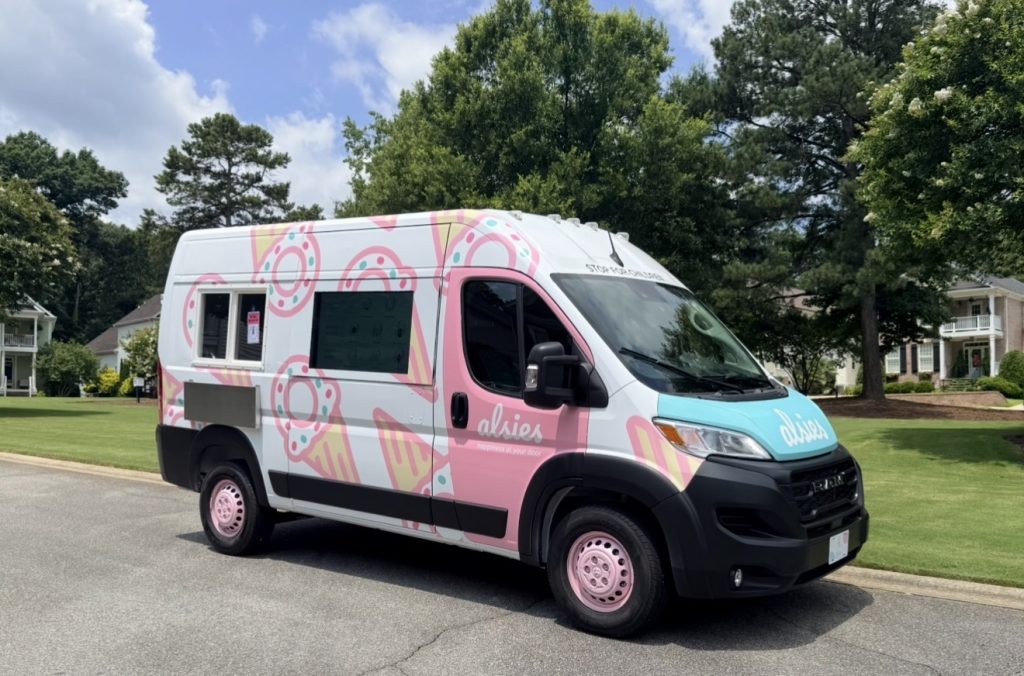 A colorful food truck with donut designs parked on a suburban street near trees and houses.