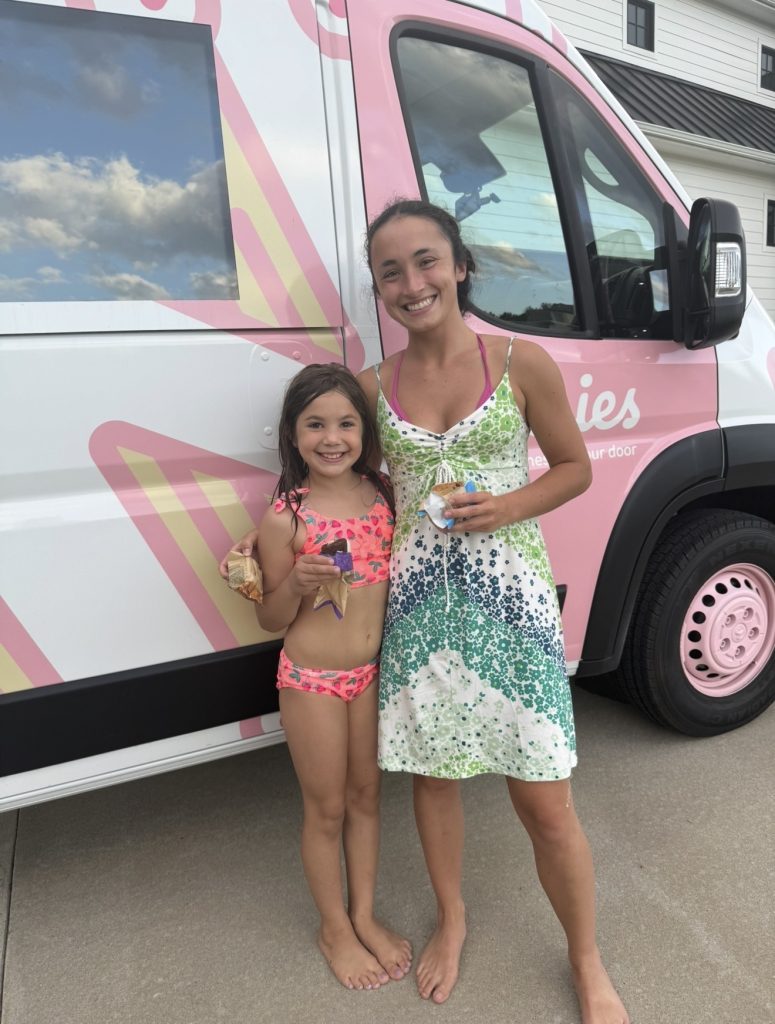 Smiling woman and girl with ice cream stand barefoot by a pink ice cream truck on a sunny day.