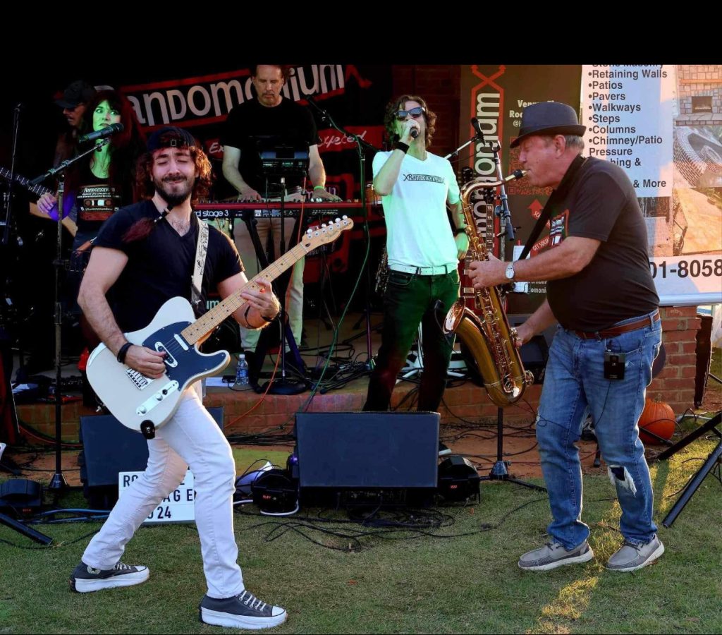 A band performs outdoors with a guitarist, keyboardist, saxophonist, and singer on a sunny day.