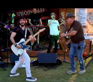 A band performs outdoors with a guitarist, keyboardist, saxophonist, and singer on a sunny day.