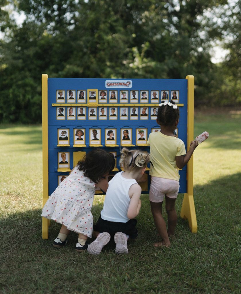 Three young children play a large outdoor Guess Who? game on grass with trees in the background.